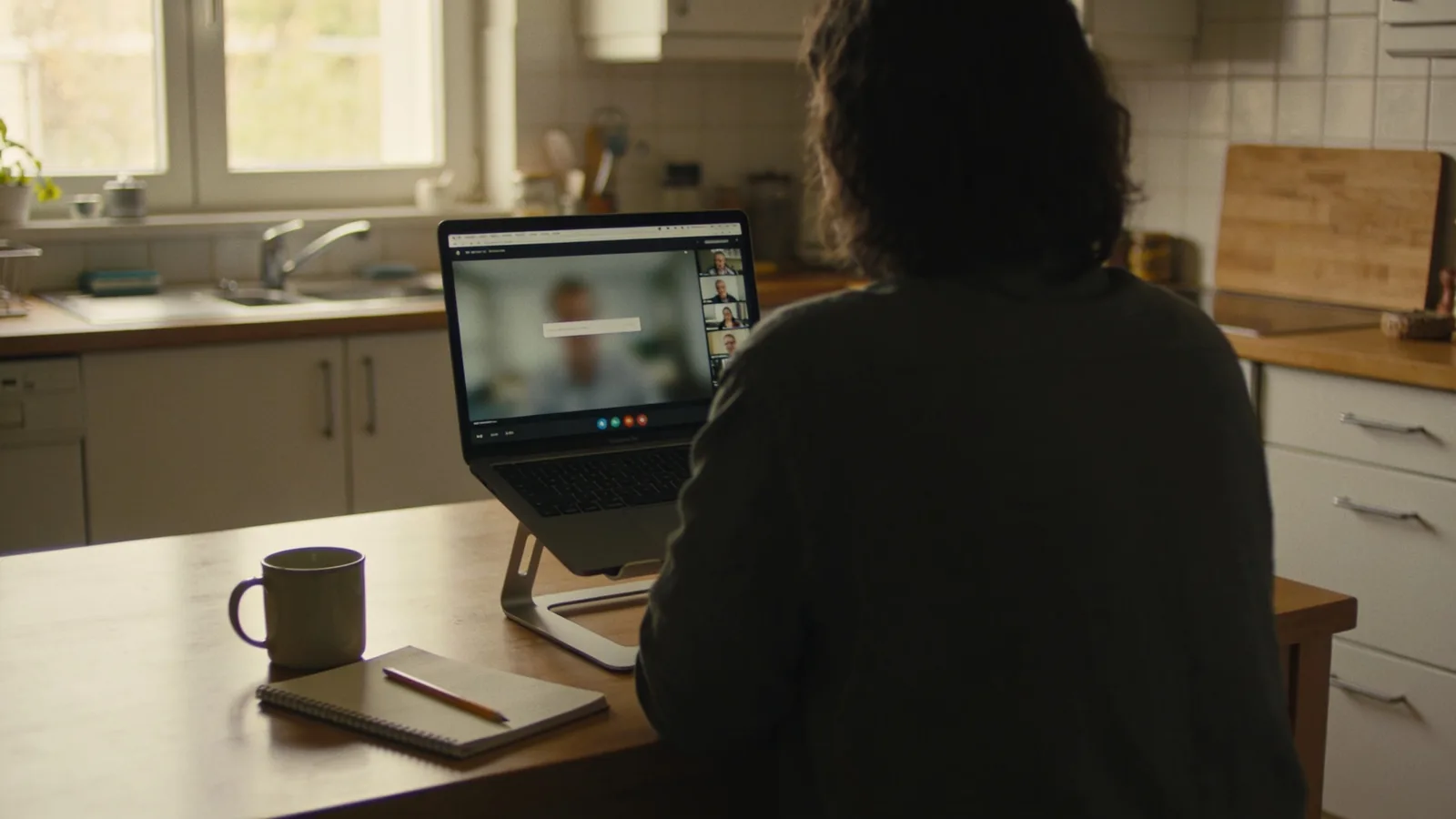 Person von hinten am Küchentisch vor einem Laptop mit Videogespräch