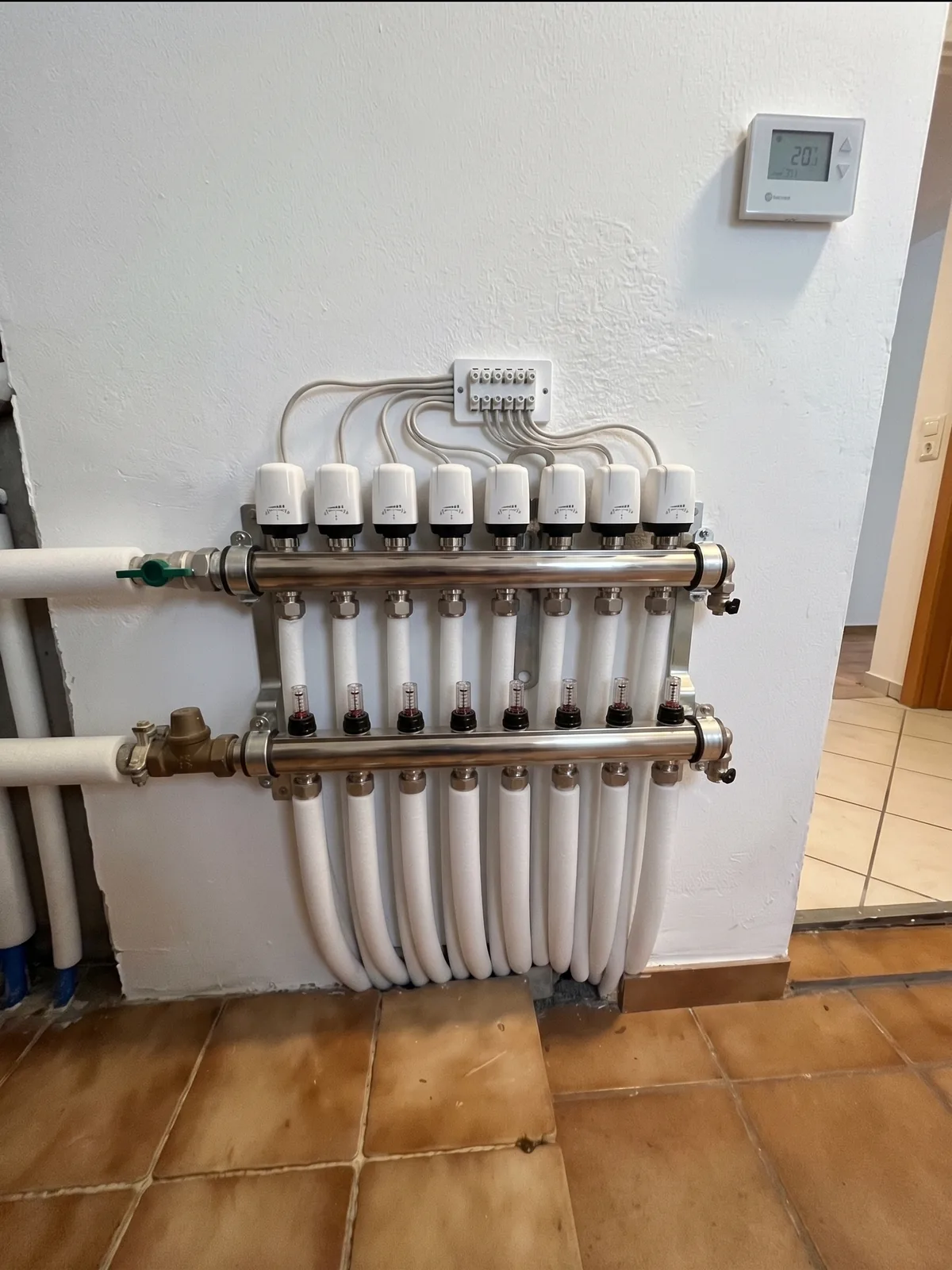Neuer Heizkreisverteiler aus Edelstahl an derselben Wand, mit weißen Ventilkappen und sauber geführten Leitungen.