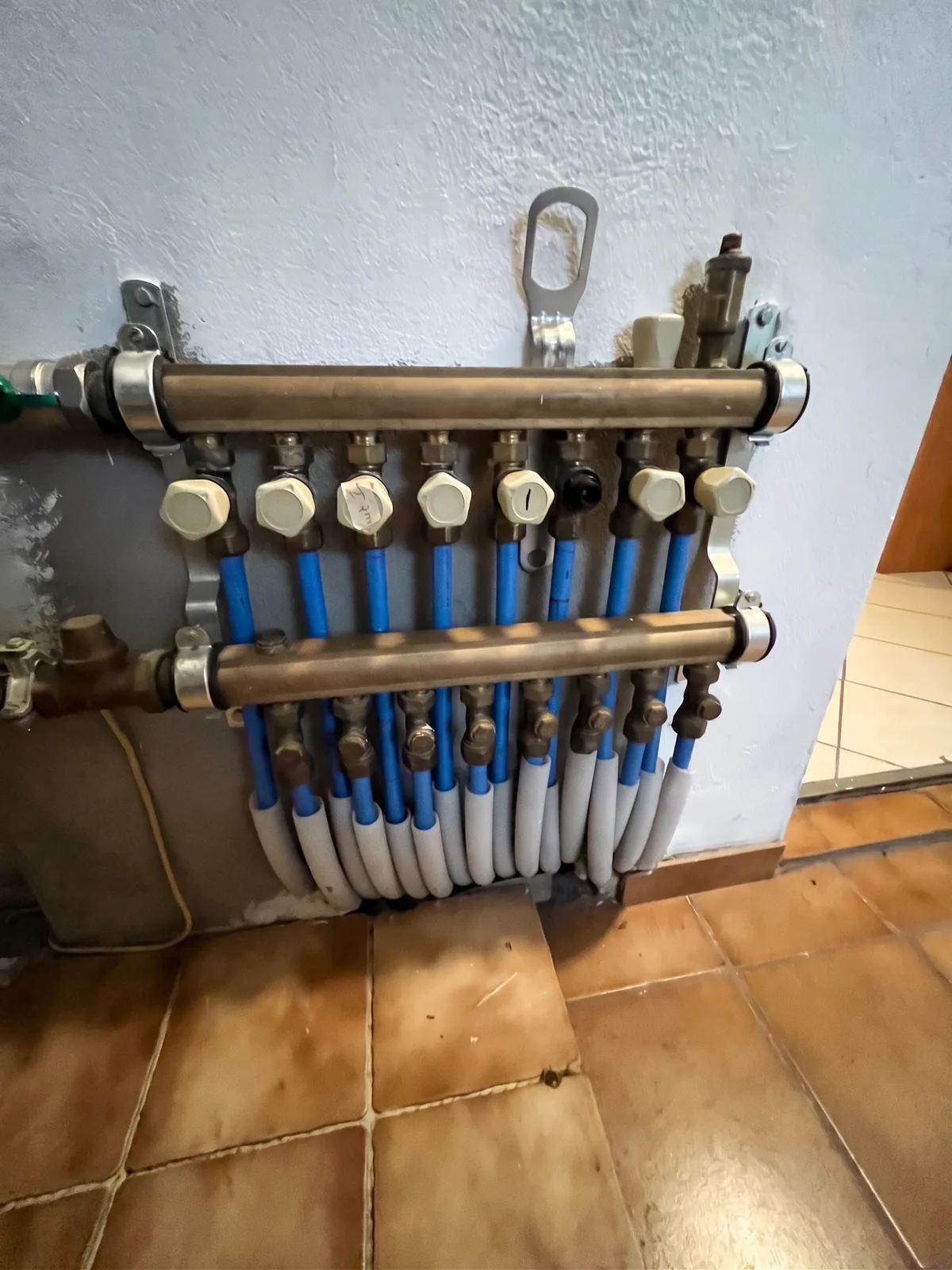 Alter Heizkreisverteiler an einer Kellerwand, mit blauen Rohrdämmungen und ungeordneten Leitungen.